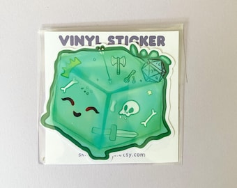 Könnte beinhalten: Ein grüner Vinyl-Aufkleber mit einem Cartoon-Würfel mit einem fröhlichen Gesicht. Der Würfel hat verschiedene spielbezogene Symbole, darunter ein Schwert, einen Schädel, einen Knochen, einen Schlüssel, eine Axt und einen 20-seitigen Würfel.