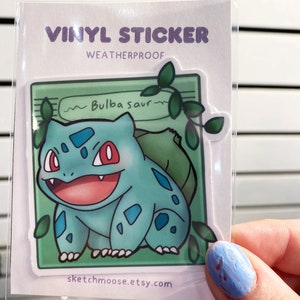 May include: A vinyl sticker featuring a cartoon Bulbasaur, a green and blue Pokemon, with a green background and vines. The sticker is weatherproof and says "~Bulbasaur~" and "sketchmoose.etsy.com".