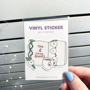 May include: A vinyl sticker featuring a cartoon illustration of a book with a cup of tea and the text "Tea & Cozy Books". The sticker is weatherproof and is available on the Etsy shop sketchmoose.etsy.com.