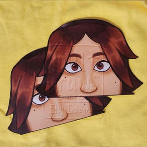 Peut inclure: Un t-shirt jaune avec un graphisme de visage aux cheveux et yeux bruns. Le visage est divisé en deux, la moitié supérieure chevauchant le bas. Les textes "raws" et "Etsy Instagram" sont visibles.