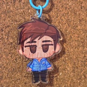 May include: A clear acrylic keychain featuring a cartoon illustration of a person with brown hair and a blue jacket. The keychain has a blue clasp and ring. The text "Instagram" is visible on the keychain.
