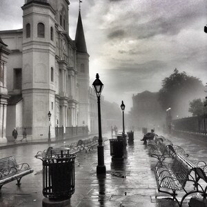 Puede incluir: Fotografía en blanco y negro de una calle de la ciudad en un día lluvioso. Una gran iglesia con campanario a la izquierda. Farolas y bancos bordean la acera mojada. Gente caminando a lo lejos.