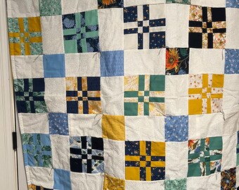 May include: A handmade quilt with a patchwork design. The quilt features a grid of squares in various colors and patterns, including floral prints, sunflowers, and geometric shapes. The squares are set against a white background.