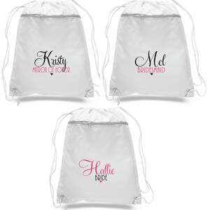 May include: Three white drawstring bags with black text and a pink heart. The bags are labeled "Kristy Matron of Honor", "Mel Bridesmaid", and "Hallie Bride".
