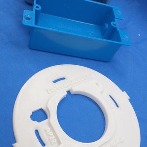 May include: A white plastic electrical box cover with the text "Parts & Technology" printed on it. The cover has a large circular opening in the center and several smaller openings around the edge. The cover is sitting on a blue surface.