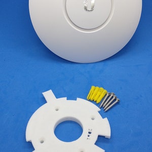 Puede incluir: Soporte de techo blanco con un logotipo U y una placa de montaje blanca con cuatro agujeros para tornillos. La placa de montaje tiene una pequeña etiqueta de texto que dice "T101E". También hay cuatro tacos de plástico amarillos y cuatro tornillos plateados.