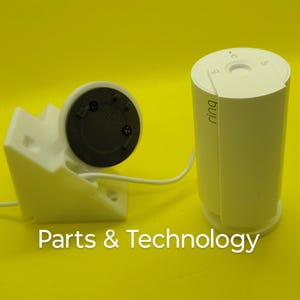 May include: A white Ring security camera with a white mounting bracket and a white power cord. The camera is connected to the bracket and the power cord is plugged into the camera. The camera is on a yellow surface.