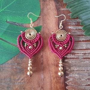 May include: A pair of maroon macrame earrings with gold wire spirals and gold beads. The earrings are hanging from gold hooks.