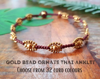 May include: A maroon cord ankle bracelet with gold bead accents. The beads are in the shape of a flower. The text "GOLD BEAD ORNATE THAI ANKLET CHOOSE FROM 32 CORD COLORS" is below the bracelet.