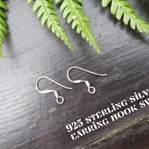 May include: Two silver earring hooks on a dark wood surface with green fern leaves. The text "925 Sterling Silver Earring Hook Swap" is printed on the surface.