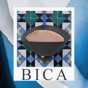 Può includere: Un portafiltro da caffè nero con un filtro di carta beige all'interno, appeso a uno spago blu su uno sfondo di piastrelle colorate. La parola "BICA" è stampata in lettere bianche sotto il portafiltro.