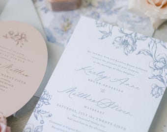 May include: Wedding invitation suite featuring a floral design in blue and white. Includes a main invitation, RSVP card, and envelope. A blush pink ring box with gold rings and fresh roses complete the elegant arrangement.