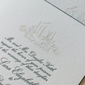 Puede incluir: Primer plano de elegantes invitaciones de boda. Las invitaciones presentan texto en relieve en una fuente de escritura, con los nombres de la pareja y los detalles del evento. El papel es blanco con un borde verde oscuro. Un monograma decorativo está en la parte superior.