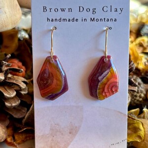 May include: A pair of dangle earrings on a white display card labeled Brown Dog Clay handmade in Montana. The earrings feature irregular, geometric-shaped pendants with a glossy, marbled finish in shades of deep purple, orange, and yellow.