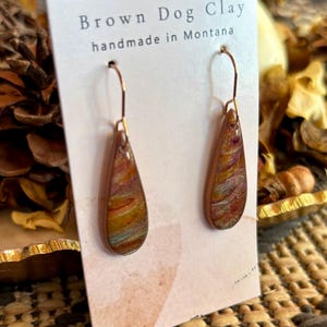 May include: Teardrop-shaped earrings featuring a marbled pattern of brown, gold, and teal tones. The earrings hang from copper-colored metal hooks and are displayed on a white card labeled Brown Dog Clay handmade in Montana.