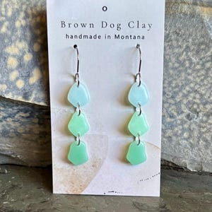 May include: Dangle earrings featuring three stacked, translucent, sea-glass-style shapes in gradient shades of light blue and green, suspended from silver-toned metal hooks on a display card labelled Brown Dog Clay handmade in Montana.