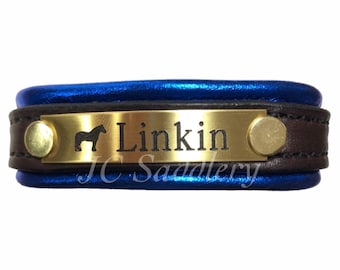 Peut inclure: Une plaque signalétique de licol de cheval en cuir bleu et marron avec le nom "Linkin" gravé sur une plaque dorée. La plaque présente également une silhouette de cheval et le texte "JC Saddlery".