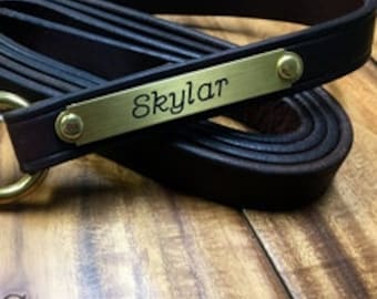 Peut inclure: Une laisse de chien en cuir marron foncé enroulée, avec un mousqueton en laiton et une plaque signalétique en laiton avec le nom "Skylar". La laisse est enroulée sur une surface en bois. Le texte "JC Saddlery" est en bas.