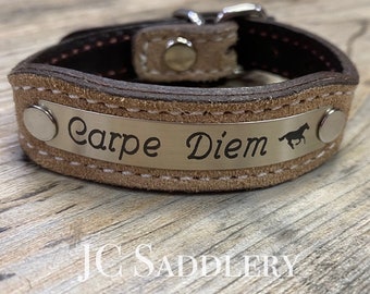 Peut inclure: Un bracelet en cuir beige avec une plaque métallique gravée « Carpe Diem » et une silhouette de cheval. L'intérieur du bracelet est marron et il est fermé par une boucle argentée. Le texte « JC Saddlery » est en bas.