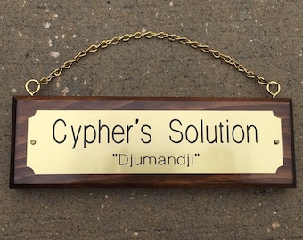 Peut inclure: Panneau rectangulaire en bois avec une plaque en laiton portant l'inscription "Cypher's Solution" et "Djumandji". Le panneau est suspendu par une chaîne dorée.