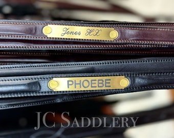 Peut inclure: Gros plan sur du matériel équestre en cuir avec des plaques signalétiques en laiton. Les plaques indiquent "Jones HX" et "PHOEBE". Le cuir est marron foncé et noir. Le logo "JC Saddlery" est visible en bas.