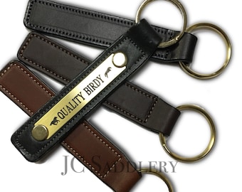 Peut inclure: Porte-clés en cuir de différentes nuances de marron et de noir, chacun avec un porte-clés doré. L'un des porte-clés comporte une plaque dorée avec le texte "QUALITY BIRDY" et une silhouette de cheval. Les porte-clés sont cousus et ont un design classique.