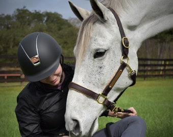 Peut inclure: Une personne portant un casque et une veste d'équitation noirs est assise sur l'herbe, interagissant avec un cheval gris. Le cheval porte un licol en cuir marron avec des accessoires dorés. La scène se déroule en extérieur par temps nuageux.