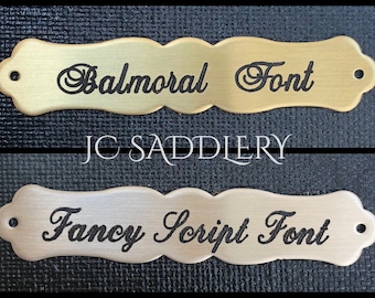 May include: Two metal nameplates with decorative edges. The top plate is gold-toned and reads "Balmoral Font" in black script. The bottom plate is silver-toned and reads "Fancy Script Font" in black script. The text "JC Saddlery" is in white script between the plates.