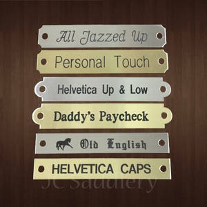 May include: A collection of engraved metal nameplates in various colors, including silver and gold. Each plate features unique text such as "All Jazzed Up" and "Personal Touch". The plates are rectangular with rounded edges and a hole on each end.