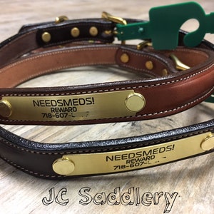 May include: Two leather dog collars in shades of brown and black, featuring brass nameplates with the text "NEEDSMEDS! REWARD" and a phone number. The collars have brass hardware and are stitched with white thread. Green tags are attached.