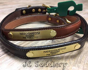 Peut inclure: Deux colliers pour chiens en cuir marron et noir, avec des plaques signalétiques en laiton portant l'inscription "NEEDSMEDS! REWARD" et un numéro de téléphone. Les colliers ont une garniture en laiton et des coutures blanches. Des étiquettes vertes sont attachées.