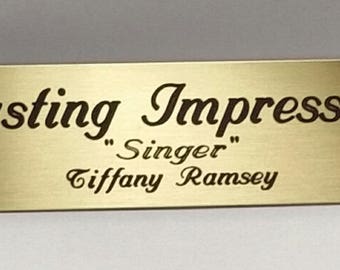Peut inclure: Plaque rectangulaire dorée avec l'inscription "Lasting Impression" dans une police décorative. En dessous, on peut lire "Singer" entre guillemets, suivi de "Tiffany Ramsey". Deux petits trous sont visibles sur le côté gauche.