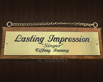 以下が含まれることがあります： 長方形の木製プラークで、金色の金属板が付いています。「Lasting Impression」という言葉と「Tiffany Ramsey」という名前が黒で刻まれています。 金色のチェーンが上部の角に取り付けられています。