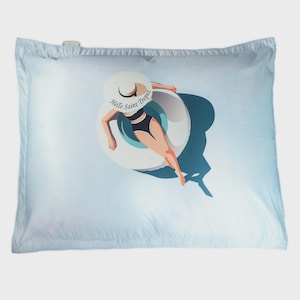 Può includere: Una federa azzurra con una grafica di una persona in costume da bagno nero e cappello bianco che galleggia su un salvagente turchese. Il cappello ha la scritta "Hello Saint Tropez". La federa ha una consistenza morbida e liscia.