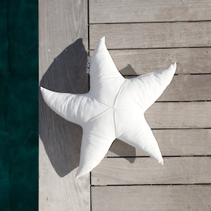 Puede incluir: Almohada blanca en forma de estrella de mar con el texto "MY HOME" en uno de los puntos. La almohada está sobre una plataforma de madera.