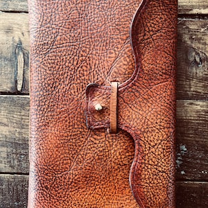 May include: A brown leather journal with a unique, wavy flap closure and a brass buckle. The leather has a textured, pebbled surface.