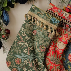 May include: Christmas stockings with floral patterns in green, red, and blue. The green stocking has a cream-colored floral design and is trimmed with gold braid and tassels. The red stocking has a floral pattern with white and yellow flowers.