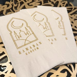 May include: Three white paper napkins with a gold design of a crescent moon, a mosque, and the text "Ramadan Kareem".