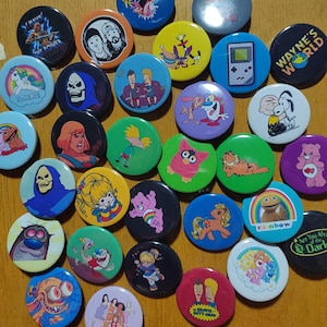 Op de afbeelding: Een verzameling van 25 kleurrijke ronde knopen met verschillende cartoonfiguren en ontwerpen. De knopen tonen personages uit populaire cartoons en shows, waaronder "Rugrats", "The Simpsons", "Ren & Stimpy", "Wayne's World", "Scooby-Doo" en "The Powerpuff Girls".
