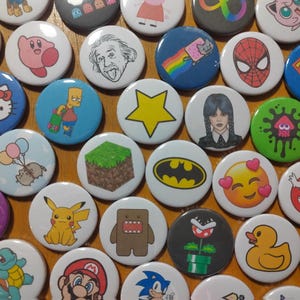 Peut inclure: Une collection de boutons ronds colorés représentant divers personnages de dessins animés, logos et motifs. Les boutons sont disposés de manière aléatoire sur une surface en bois. Parmi les personnages, on trouve Hello Kitty, Pikachu, Bob l'éponge et un bloc Minecraft.