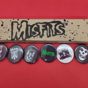 May include: A collection of six round Misfits band buttons with various skull and logo designs. The buttons are black, white, and red, with a cardboard box featuring the band's name in black. The background is red.