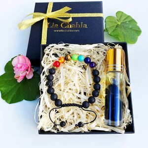 Könnte beinhalten: Eine schwarze Geschenkbox mit goldenem Band enthält ein Chakra-Armband und eine Glasflasche mit ätherischem Öl. Das Armband hat schwarze Lavaperlen und farbige Edelsteinakzente. Die Flasche hat einen goldenen Deckel und enthält eine blaue Flüssigkeit.