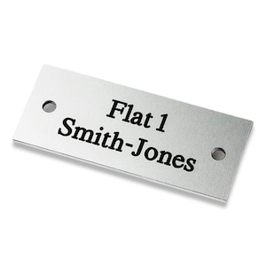 May include: Silver metal door sign with black text that reads "Flat 1 Smith-Jones".