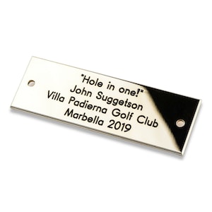 May include: A gold-colored metal plaque with the text "Hole in one!" John Suggetson Villa Padierna Golf Club Marbella 2019.