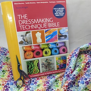 Puede incluir: Un libro amarillo titulado "The Dressmaking Technique Bible" de Lorna Knight. La portada del libro presenta un maniquí de costura, carretes de hilo, botones y una cremallera. El libro está sobre una tela blanca con un patrón de corazones arcoíris.