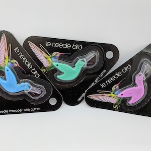 May include: Three needle threaders shaped like hummingbirds, each packaged in a triangular card with a hummingbird illustration. The threaders are blue, green, and pink.
