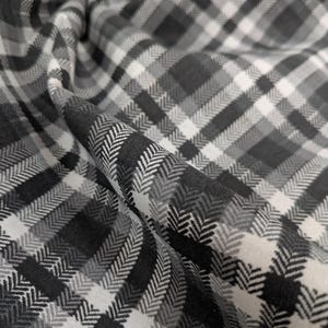 May include: Close-up of a fabric with a black, white, and grey plaid pattern. The fabric has a herringbone weave, creating a textured appearance. The fabric is folded, showing the pattern's direction and depth.