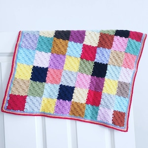 Op de afbeelding: Een kleurrijk gebreid babydekentje met een patchwork ontwerp. Het dekentje is gemaakt van vierkanten in verschillende kleuren, waaronder rood, blauw, groen, geel en paars.