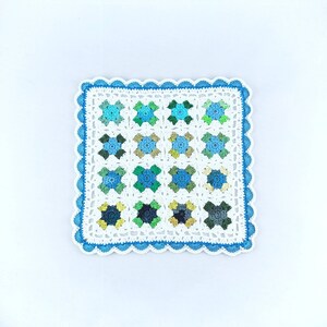 May include: A white crocheted square with a blue border and a scalloped edge. The square is made up of smaller squares in shades of blue, green, and yellow.