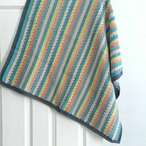 Puede incluir: Una manta de ganchillo multicolor con rayas verticales en tonos pastel de azul, rosa, amarillo y beige. La manta tiene un borde gris y est&aacute; colgada sobre una puerta blanca. El patr&oacute;n de ganchillo presenta un dise&ntilde;o de puntada abierta y texturizada.